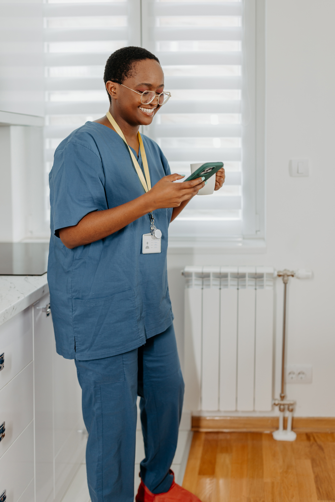 A young African American female nurse is holding a cup of coffee and cheerfully looking at her phone.